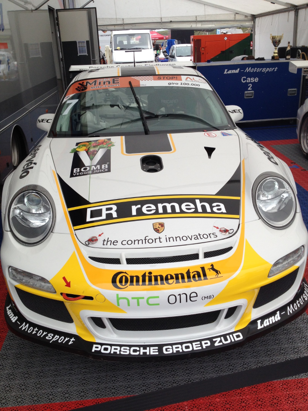 Weißer und gelber Porsche 911 GT3 RSR Rennauto in einer Garage geparkt mit anderen Fahrzeugen, Menschen und Gegenständen im Hintergrund.