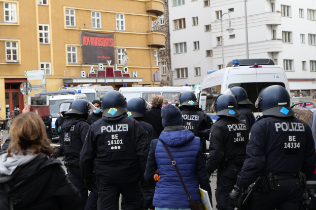 Polizeibeamte in Uniform vor einer Menge mit Helmen und Jacken, mit Fahrzeugen, Gebäuden, Laternenmasten und einem Banner im Hintergrund während einer Demonstration in Berlin, Deutschland.