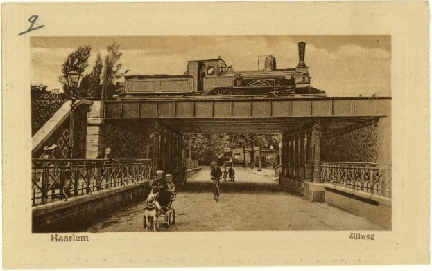Schwarz-weiß-Foto einer Bahnübergang über eine Brücke mit Menschen auf Fahrrädern und einer Person in einem Kinderwagen, Bäume im Hintergrund und Text unten.