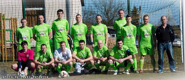 Gruppe von Männer in grünnen T-Shirts auf einem Fuballfeld mit einem Ball und einem Torpfosten, Bäumen und Gebäuden im Hintergrund.