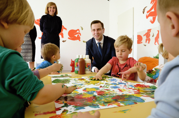 Kinder sind in einer Malaktivität um einen Tisch mit bunten Händen, Papieren und Flaschen versammelt, die von einer lächelnden Frau beaufsichtigt wird, mit Kunstwerken an der Wand im Hintergrund.